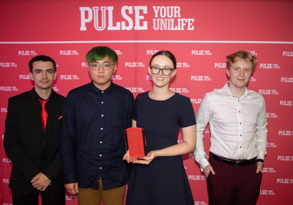 4 people, 1 holding a red award in front of a red and white backdrop